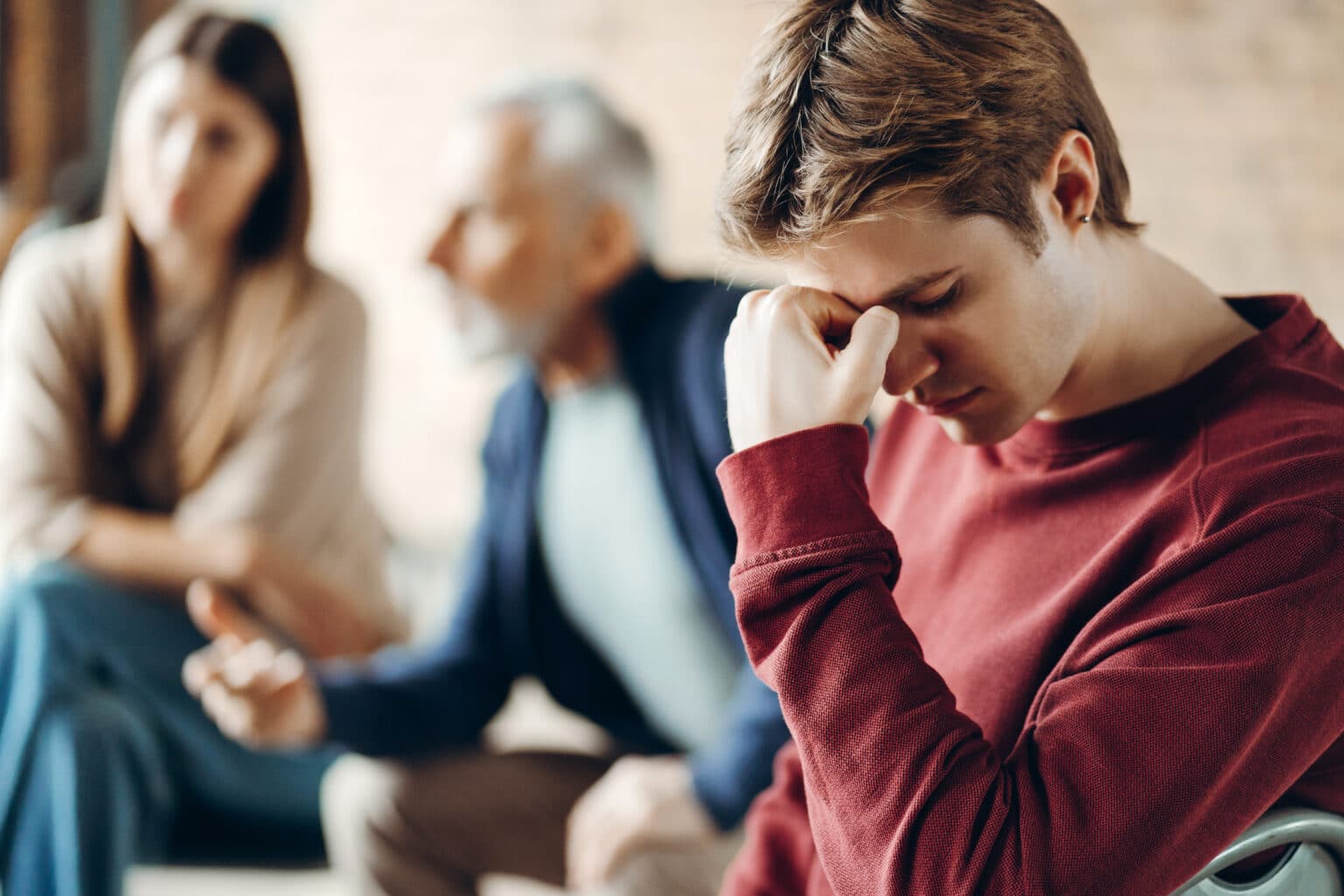A young man with his head down while his parents talk to someone in the background for the topic how to choose a rehab for your son.